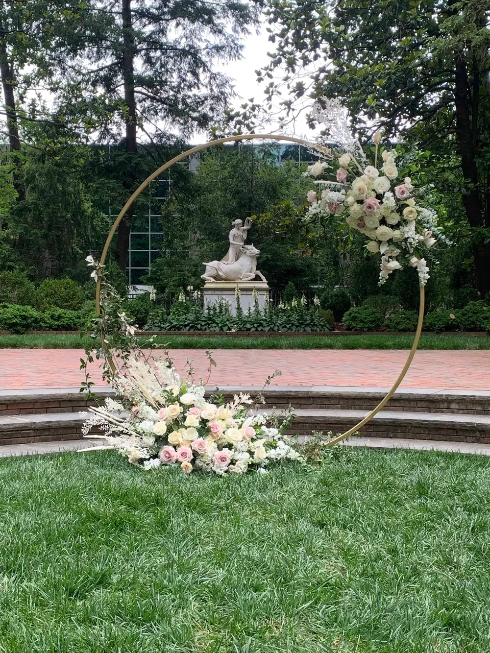 Circular ceremony arch with blush and white florals in front of a garden statue